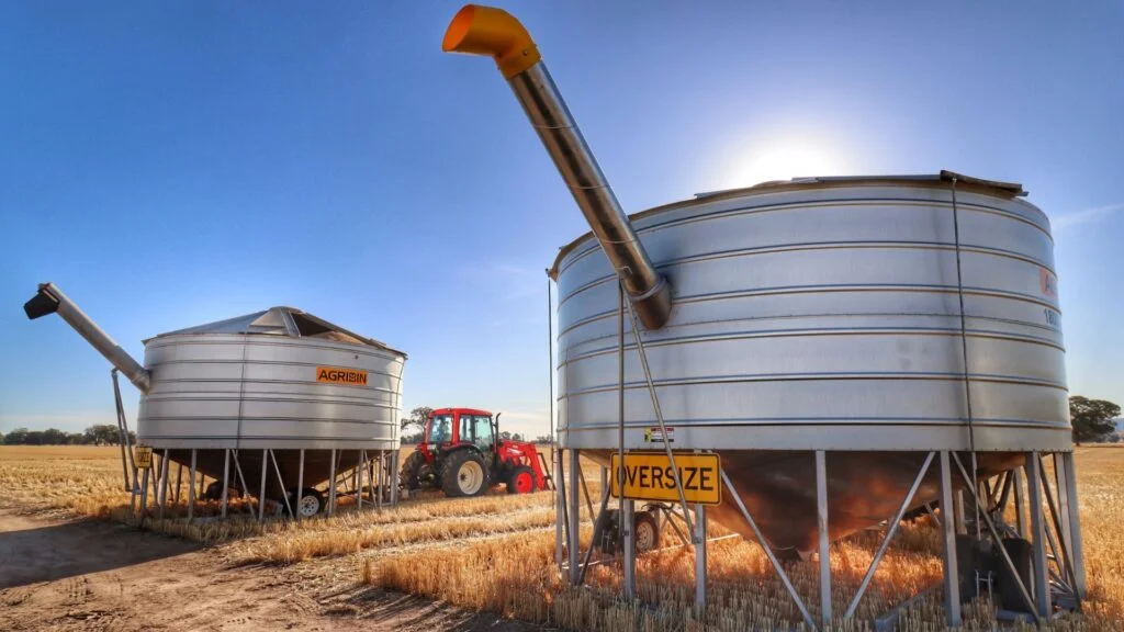 Two AGRIBIN field bins standing in a stubble paddock with a tractor between them, augers raised